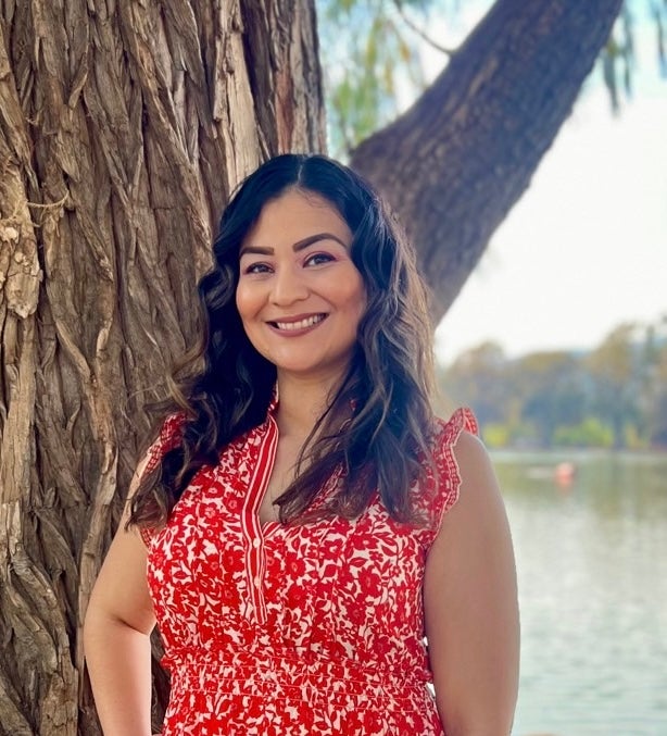 Photo of Monica Cardenas in a red and white dress, standing in front of a tree and lake.