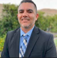 Photo of Ruben Fierro with a dark grey suit jacket, blue dress shirt and a grey, blue and white multicolored tie.